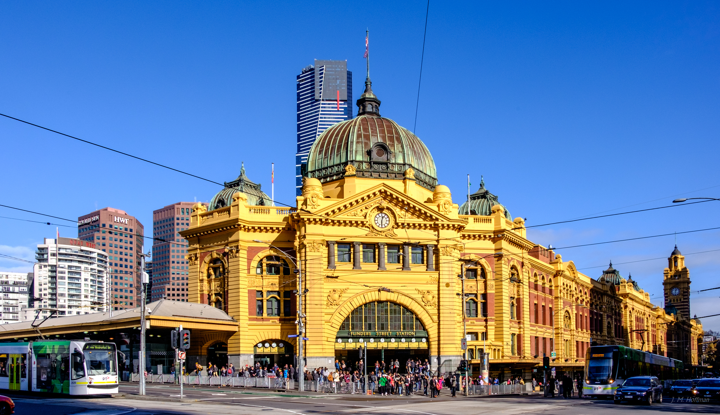 a large yellow building with a dome and a crowd of people in front with Flinders Street railway station in the background