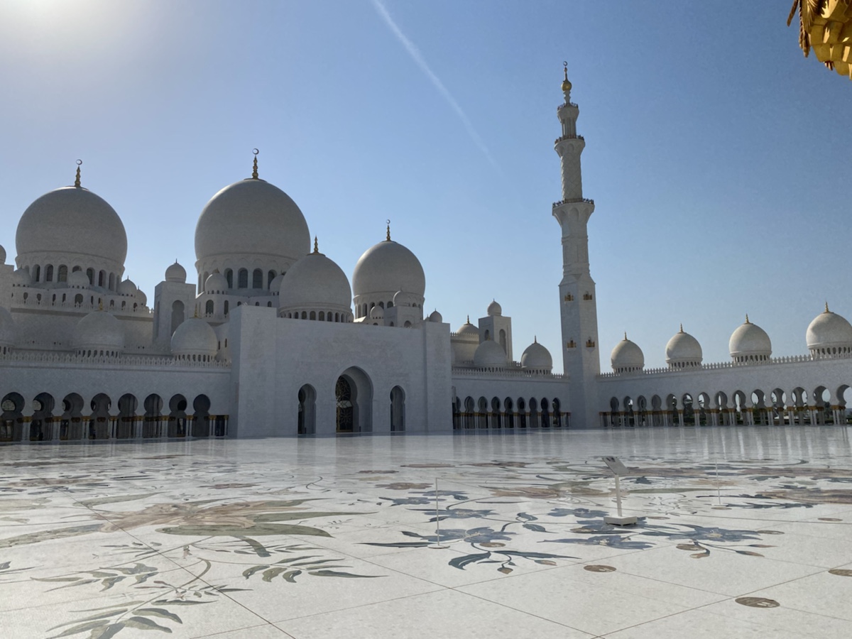 a large white building with domes and a tall tower with Sheikh Zayed Mosque in the background