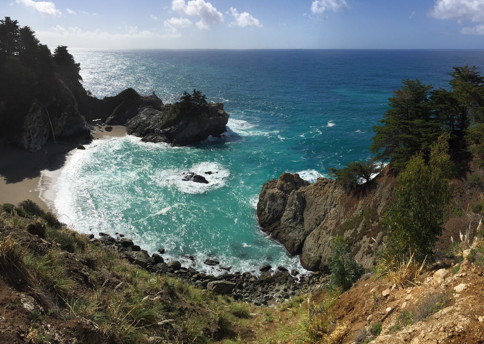 a rocky beach with trees and a rocky cliff with McWay Falls in the background
