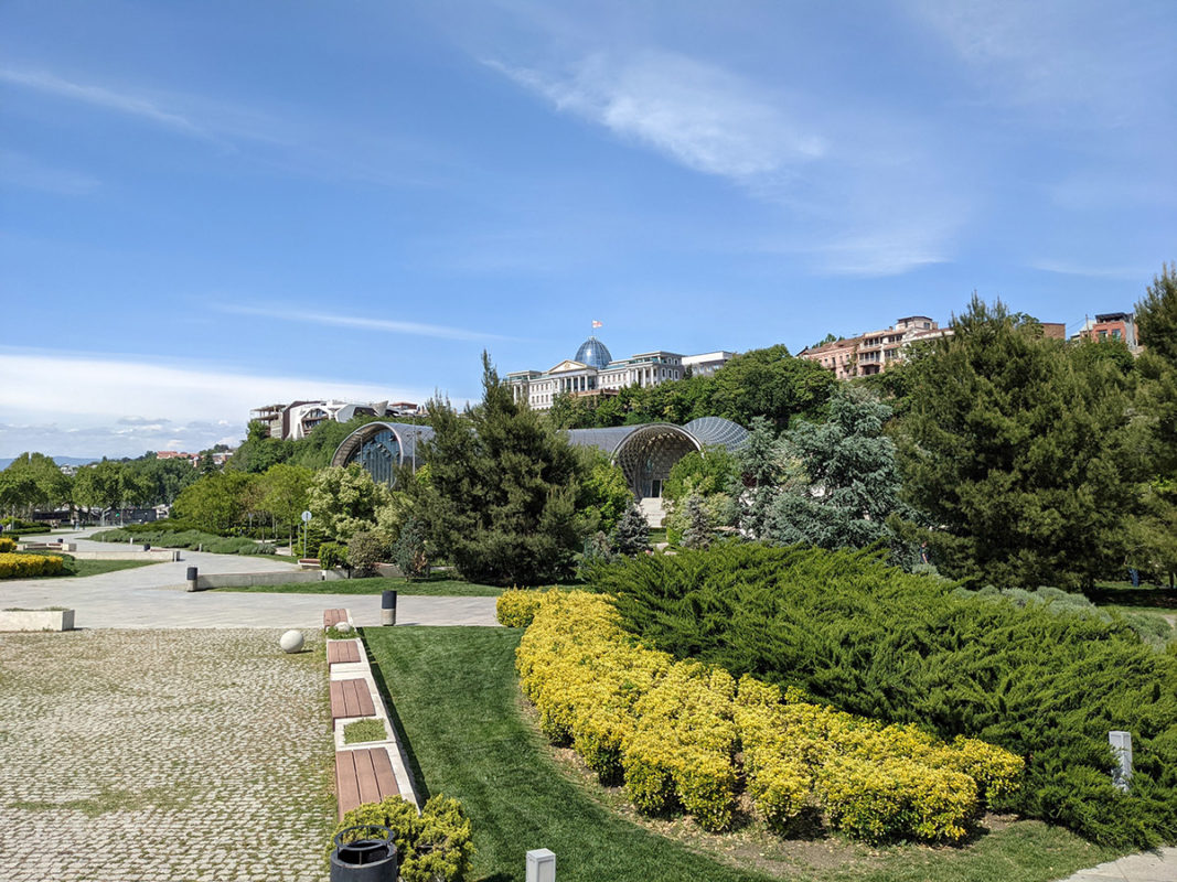 a park with a stone path and bushes and buildings in the background