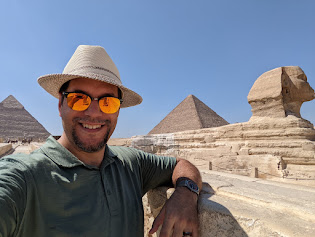a man in a hat and sunglasses leaning on a stone wall with pyramids in the background