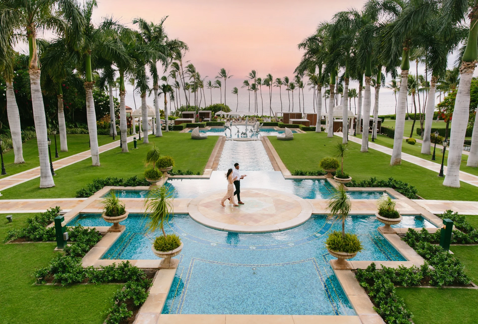 a man and woman standing in a pool with palm trees and a pink sky